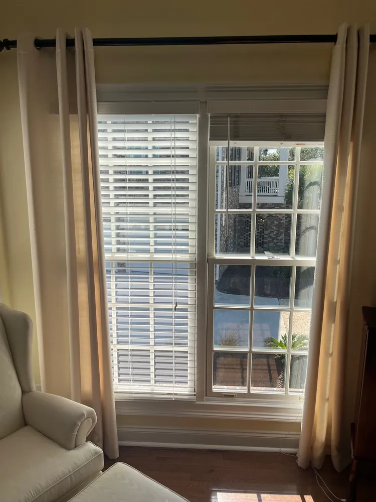 Original paired windows with blinds at different heights, light curtains and a chair beside the opening